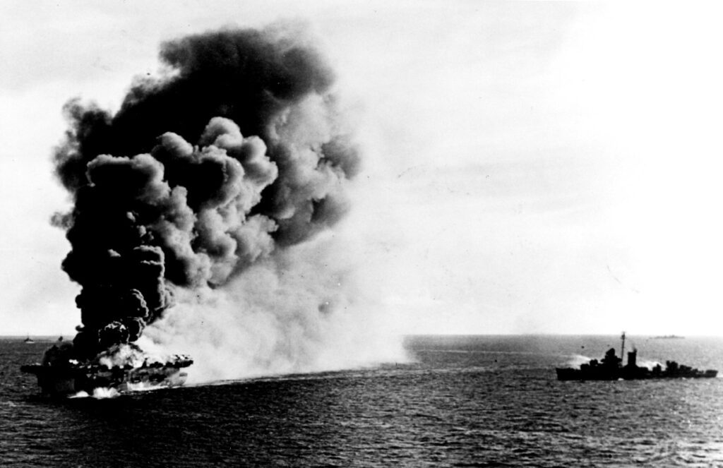 The burning carrier, with a destroyer standing by with fire-hoses (Robert O Baumrucker / NHHC)