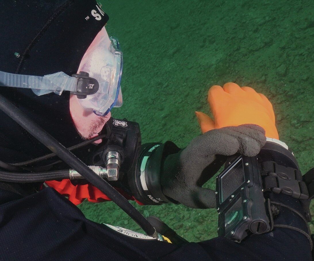 Ratio iX3M 2 GPS Tech+ Dive Computer Review: Multi-Gas Colour Dive Computer with GPS and Wireless Air Integration 4 Diver wearing a drysuit viewing Ratio iX3M 2 GPS Tech+ dive computer underwater, showing large colour display.
