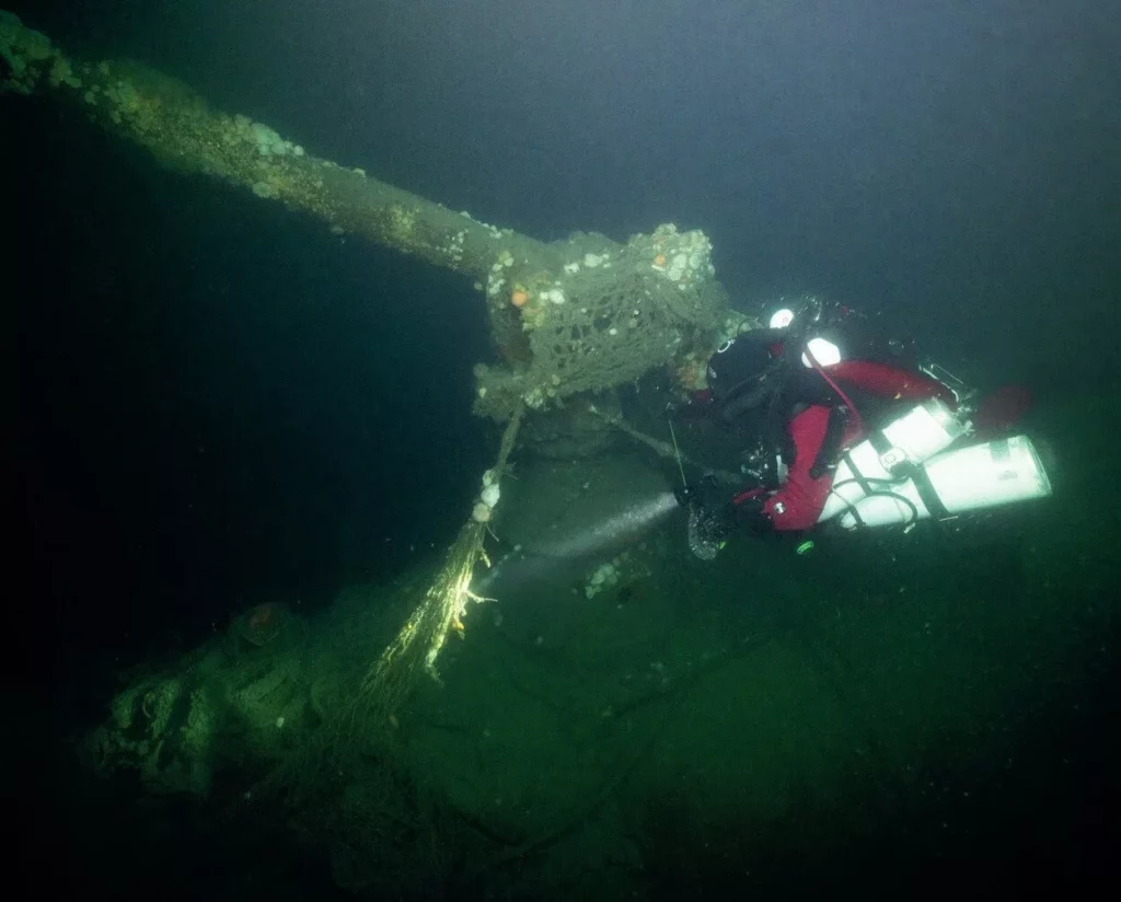 A technical diver looks at the large 105mm gun of the U58 pointing at a 45-degree angle from the wreck hull.