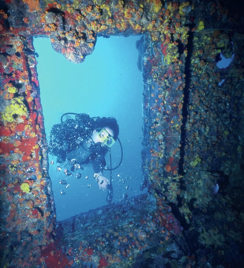 Scuba divers swimming through the open iron ribs and interior structure of the RMS Rhone wreck.
