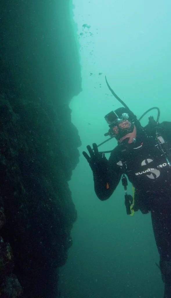 A scuba diver drifting alongside a dramatic underwater wall at Big Geiger Cove, Catalina Island, surrounded by deep crevices and marine life.