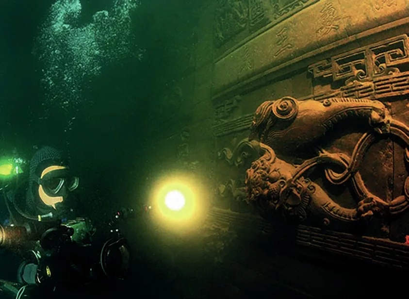 A scuba diver with a torch exploring the ancient stone walls and ornate carvings of the submerged Lion City ruins.