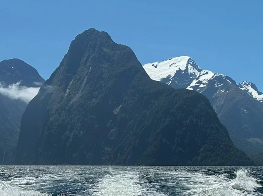 Panoramic view of a deep blue fjord in Fiordland, New Zealand, with steep snow-capped mountains and lush green rainforest cliffs reflecting in the water.
