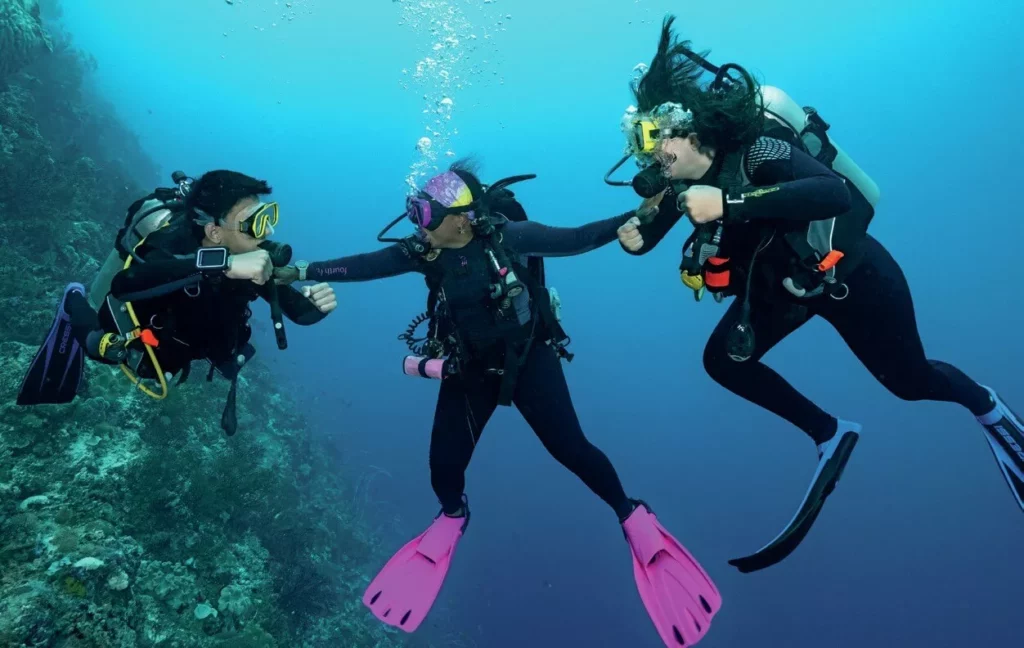 Aura and Ian Chatham giving enthusiastic signs underwater near a healthy coral reef wall while scuba diving in Wakatobi.