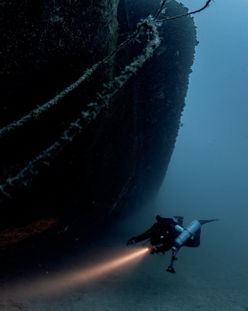 Diver exploring the exterior of the HMS Southwold wreck in clear Mediterranean waters
